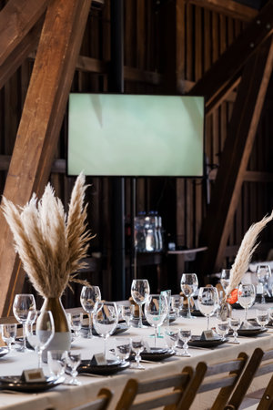 An elegantly set wedding table inside a rustic barn features white linens, black plates, glassware, pampas grass arrangements, and a projection screen, creating a romantic farm venue atmosphereの写真素材