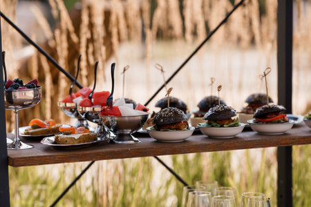 An elegant outdoor catering display features mini black burgers, fresh fruit skewers, and assorted appetizers presented on a wooden shelf, ideal for events and celebrationsの写真素材
