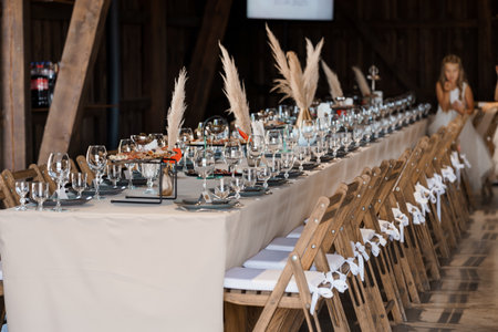 A long wedding reception table is beautifully set inside a rustic barn, featuring beige linens, wooden chairs with white ribbons, and pampas grass centerpieces. an elegant, warm mood prevailsの写真素材