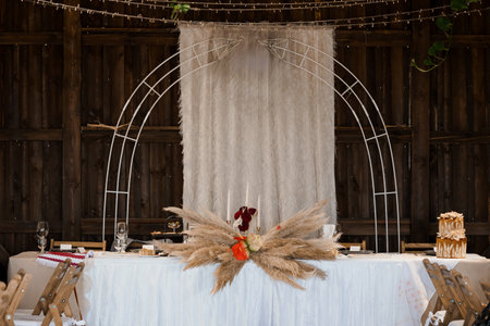 A beautifully styled wedding head table features a metal arch draped with sheer fabric and adorned with pampas grass. candles and floral arrangements create a romantic barn reception moodの写真素材