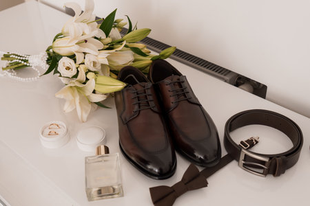 Stylish groom's wedding preparation flat lay featuring dark brown leather shoes, matching bow tie, pearl necklace, perfume bottle, and belt on a pristine white surface, conveying luxuryの写真素材
