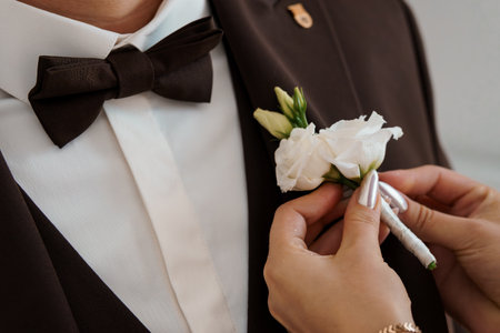 A close-up depicts shots of a person meticulously pinning a delicate white flower boutonniere onto the lapel of a grooms dark brown tuxedo, showcasing a romantic wedding momentの写真素材