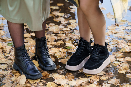 Autumn Fashion Boots Two Womens Legs and Black Leather Ankle Boots Standing in Fallen Leaves, Outdoor Style, Fall Colors, Close-Up Footwear.の写真素材
