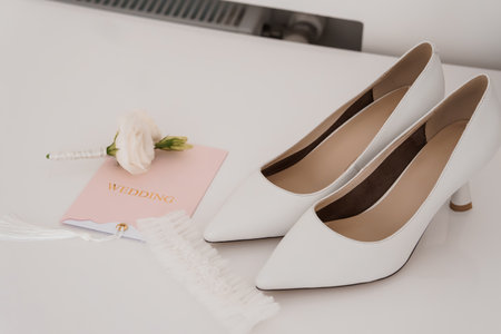 Elegant White Wedding Shoes with Rose Detail and Invitation Card on a Light Surface, Stylish Bridal Accessories, Romantic Celebration Theme.の写真素材