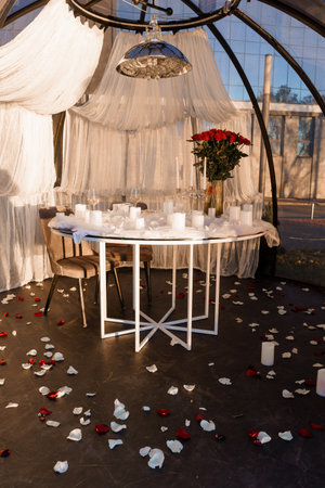 Intimate dining scene inside a greenhouse with a round table adorned with white candles, red roses, and scattered petals, creating a romantic ambiance for a couples special occasionの写真素材