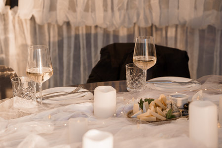 An intimate dinner for two features wine glasses filled with a light-colored beverage, a cheese plate, and soft candlelight, set against a backdrop of flowing white drapery, evoking romanceの写真素材