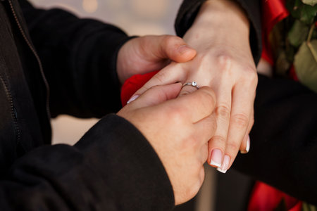 A tender engagement scene captures a couples hands as a sparkling diamond ring is presented, framed by vibrant red roses during a sunset proposal. symbolizing love, commitment, and joyの写真素材
