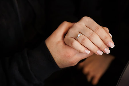 A tender close-up depicts a couple's hands clasped together, showing a sparkling diamond engagement ring on the woman's finger. the dark clothing creates an intimate and modern moodの写真素材