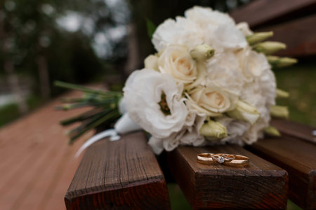 A close-up of a white wedding bouquet with roses and lisianthus resting beside gold wedding rings on...