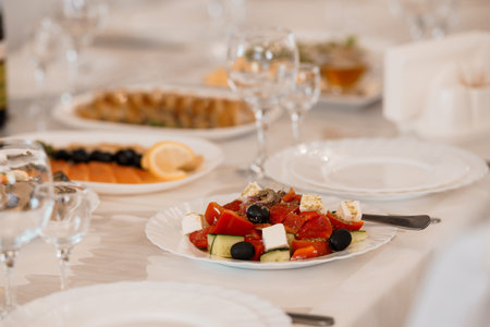A beautifully arranged Greek salad with vibrant red tomatoes, green cucumbers, black olives, and white feta cheese sits on a white plate, surrounded by wine glasses and other dishes at an upscale restaurant settingの写真素材