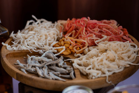 A beautifully arranged wooden platter displays an assortment of dried seafood including white fish strands, vibrant red shrimp, golden peanuts, and small silver fish, ideal for culinary or cultural food presentationsの写真素材