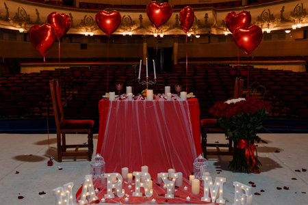 A beautifully arranged romantic proposal scene with heart-shaped red balloons, candles, and roses on a table in an elegant theater, perfect for engagement or Valentine's Day celebrationsの写真素材