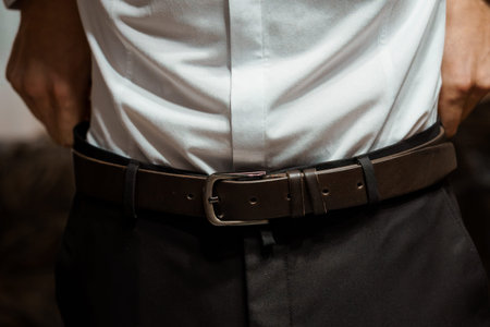 A stylish close-up of a man adjusting his brown leather belt over a crisp white dress shirt,...