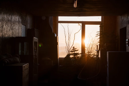 Dark room interior with dramatic backlighting from a golden sunset through a large window,...