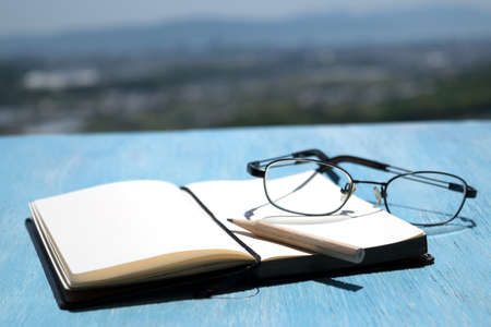 Open notepad with glasses on a wooden table with pencil on background blue sky and mountainの写真素材