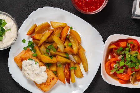 Fried fish and chips on a white plate on black the kitchen table with tomato sauce, tartar sauce and pickled vegetables salad - photo, imageの写真素材