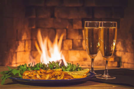 Two glasses with champagne and a plate with shrimps, lemon and greens on a wooden table against the background of a fireplaceの写真素材