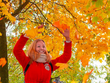 SAINT-PETERSBURG, RUSSIA - OCTOBER 8, 2018: Young beautiful girl blonde in a red jacket and her boyfriend enjoying a warm autumn day in the park and having fun playing with autumn maple leavesのeditorial素材