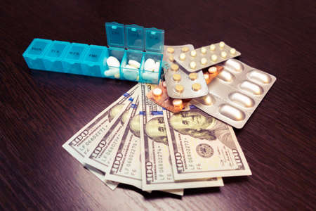 Pillbox, pills and tablets on dollar money on dark wooden table. Medicine expenses. High costs of...