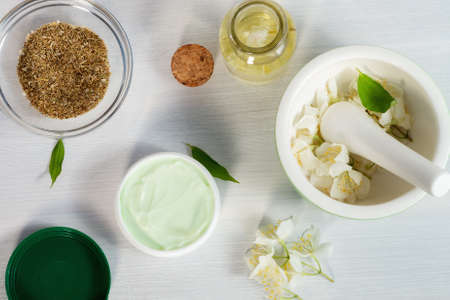 Jar of cream made from natural ingredients, oils and herbs, jasmine flowers, mortar and pestle on a white wooden background - preparation of organic cosmetics concept, close up, top view, flat layの写真素材