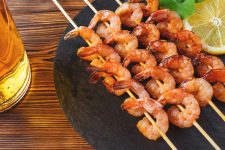Shrimp fried on skewers with arugula, lemon on a black slate dish and beer in a glass on a wooden tableの写真素材
