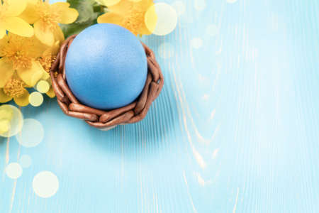 One blue egg in a wicker stand on a blue wooden table with yellow flowers - blank for design or greeting card, place for text, copy space, top viewの写真素材