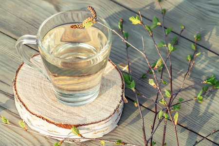 Birch juice on a wooden table in a glass mug, next to a branch of birch with young leavesの写真素材
