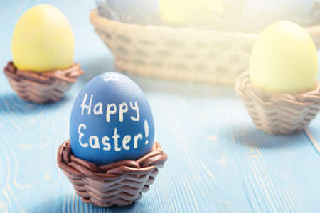 Easter greeting card - blue egg in a wicker stand with the inscription Happy Easter and a basket with colored eggs on a blue wooden table.の写真素材