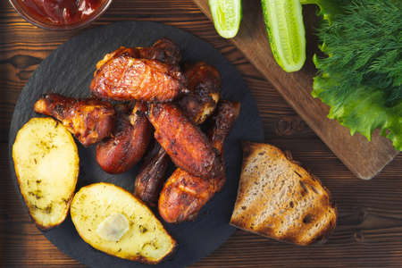 Grilled chicken drumsticks and wings with fresh cucumbers, tomato sauce, baked potatoes and herbs on a wooden table, top view, flat layの写真素材