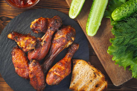 Grilled chicken drumsticks and wings on a black slate dish with fresh cucumbers, tomato sauce and herbs, top view, flat layの写真素材