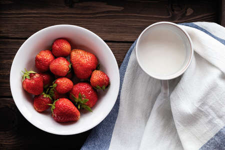 Natural ripe strawberries in a white bowl and milk in a mug on a dark wooden background. Top viewの写真素材