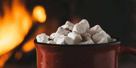 Cocoa with marshmallows and chocolate in a red mug near a burning fireplace, horizontal bannerの写真素材
