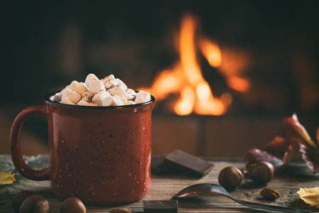 Cocoa with marshmallows and chocolate in a red mug on a wooden table near a burning fireplaceの写真素材