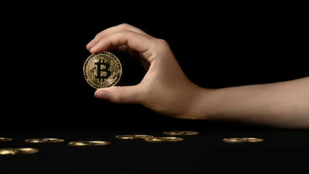 Womans hand holds a bitcoin over a black table on which other coins lieの写真素材