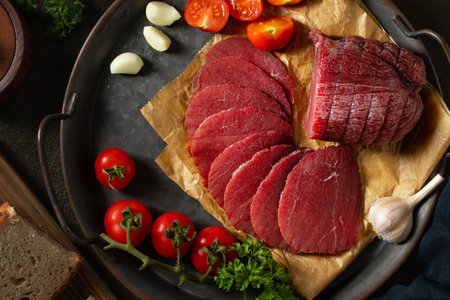 Whole and sliced bresaola on a metal round tray with tomatoes, garlic and bread