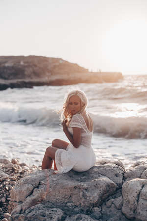 Lonely beautiful romantic girl with blonde wavy hair in white sexy dress looking at the sunset and storm on the sea and dreaming. Freedom conceptの写真素材