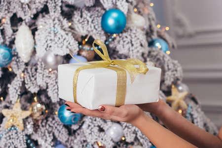 Female hands holding gift box decorated with golden ribbon, on Christmas tree background