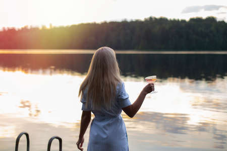 Young woman with blonde hair holding glass of champagne on lake background at sunset timeの写真素材