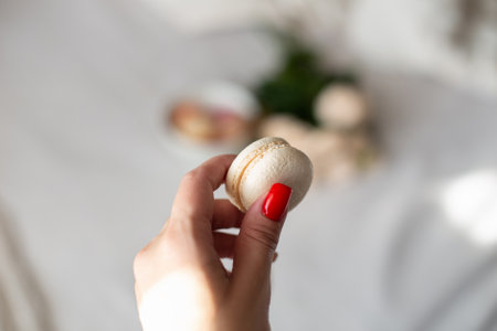 Female hand with red nails holding vanilla macaroon on white roses backgroundの写真素材