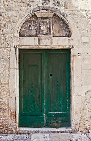 Ancient portal with a green wooden door encased in a white stone wall, surmounted by a marble arch with a carved shield;のeditorial素材