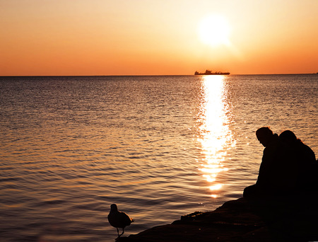 Couple sitting on a pier with seagull silhouette, sunset sunlight on sea waters and red sky
