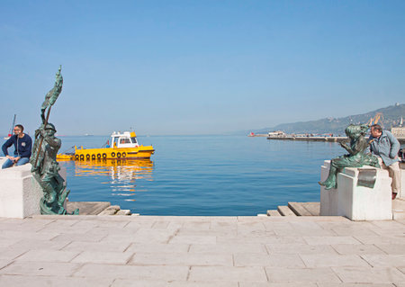 Trieste, Italy - waterfront sea promenade Le Rive with an antipollution boat crossing by.Pier with bronze monuments to the side of the landing stage, an Italian infantryman bersagliere to the left and two Trieste girls to the right.のeditorial素材