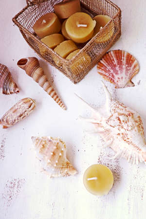 Still life: beeswax candles in a gold wire mesh box with sea shells on a white wooden table, top...