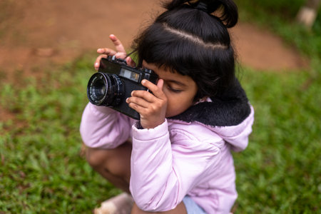 A young girl with pigtails and a big smile holds a DSLR camera up to her eye. She is focused on taking a picture of something in the distance. The background is a lush greenの写真素材