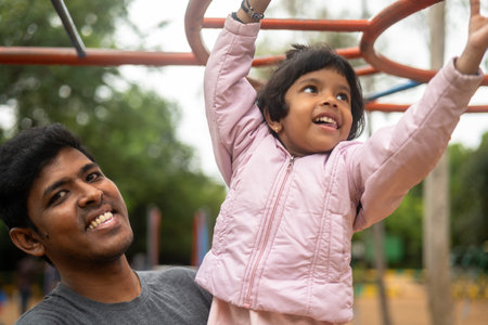 A father and daughter are climbing monkey bars in a park. The father is helping his daughter reach...