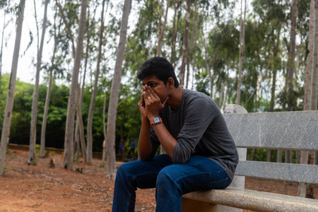 photo of a depressed Indian man sitting on a bench in a park. He is looking down at the ground with...