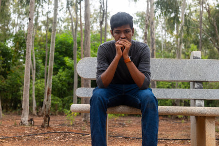sad Indian man sitting on a bench in a park. He is looking down at his hands, and his expression is one of deep despair.の写真素材