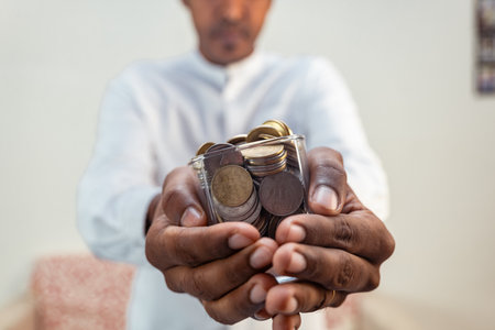 A man holds a glass filled with Indian rupees coins. The coins are of various denominations and colorsの写真素材