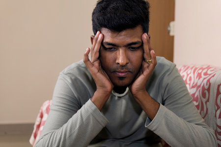 young sad and depressed Indian man suffering depression problem at home living room sofa couch...