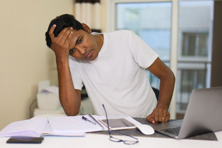 young stressed and worried Indian man working on a laptop computer at home office desk feeling...